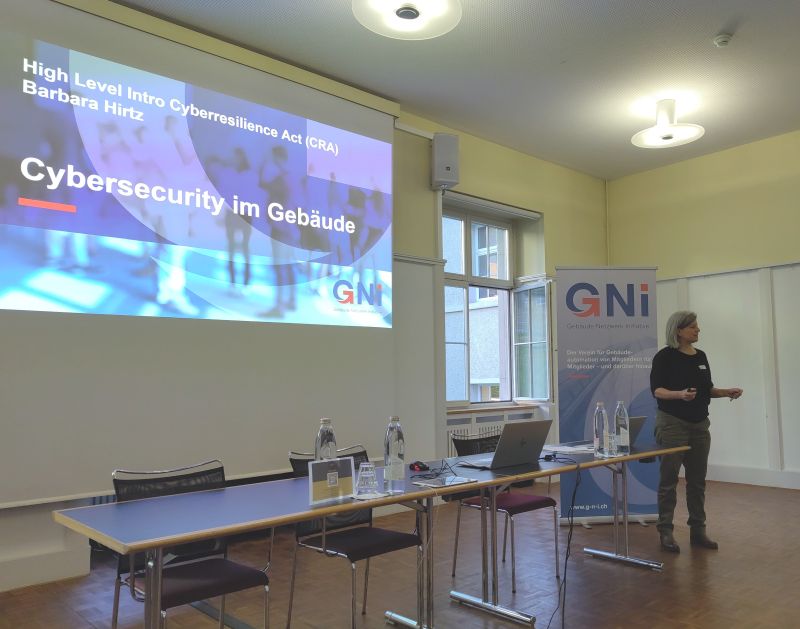 Eine dunkel gekleidete Damen steht vor einer Leinwand, auf der eine blaue Präsentation gezeigt wird.